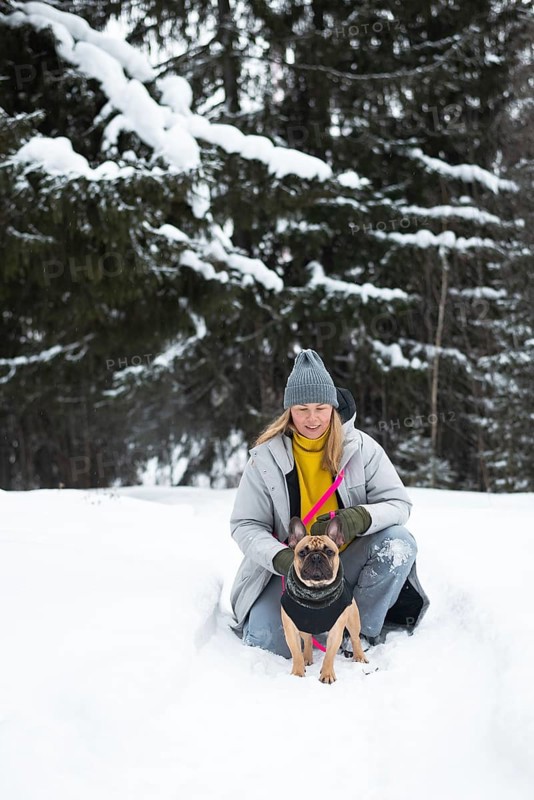 A woman in a winter coat kneels with a dog in a snow-covered forest. The serene winter landscape adds charm to this peaceful pet outing in nature