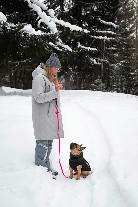 A woman in a warm coat and hat walks her dog on a snowy trail. The forest around is covered in snow, emphasizing the serene winter atmosphere