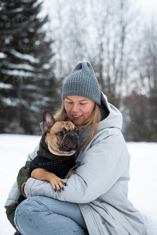 A woman in winter attire lovingly holds her French Bulldog in a snowy landscape. The scene captures the warmth and companionship between them amidst the cold surroundings