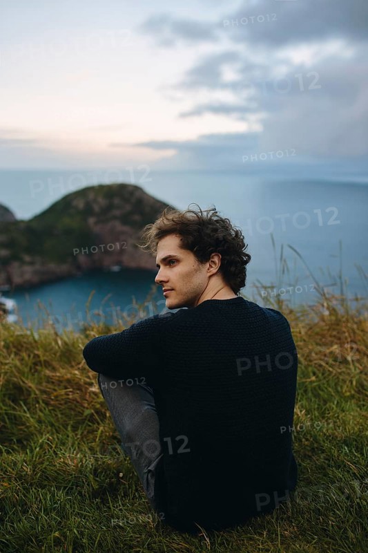 A young male sits serenely overlooking the calm waters from Signal Hill, St. John's. His relaxed pose contrasts with the wild, natural setting around him, capturing a moment of peace