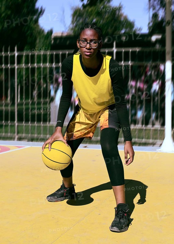 A focused woman basketball player dribbles a ball on a vibrant outdoor court. Dressed in a yellow jersey, they practice their skills under bright sunlight, embodying athleticism
