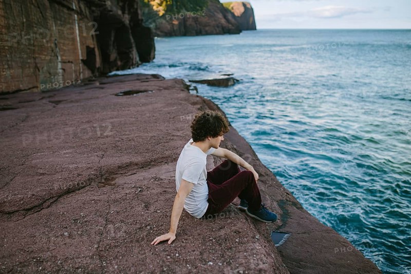 A young male sits contemplatively by the serene sea at Flatrock, Newfoundland, enjoying a tranquil afternoon amidst natural surroundings