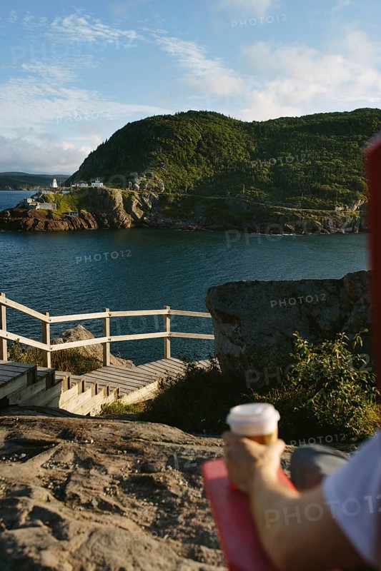 Cropped unrecognizable person sitting on rocks at Signal Hill, holding a coffee cup with a scenic overlook of the Fort Amherst in the rugged coastline and green hills of St. John's in Newfoundland & Labrador, Canada