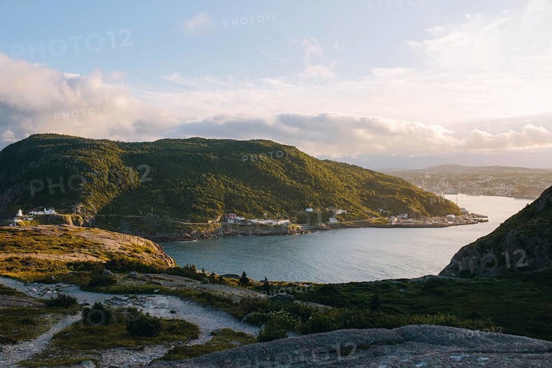A serene view of Signal Hill, St. John's showcasing a sweeping coastline with a sunlit harbor nestled between lush, green hills during the golden hour in Newfoundland & Labrador, Canada