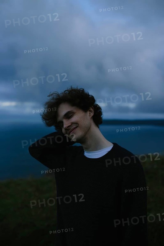 A young man stands with eyes closed relaxed against a backdrop of a cloudy sky and distant sea at Signal Hill in St. John's, Newfoundland & Labrador, Canada. He is smiling gently, with his hand running through his hair in a contemplative moment