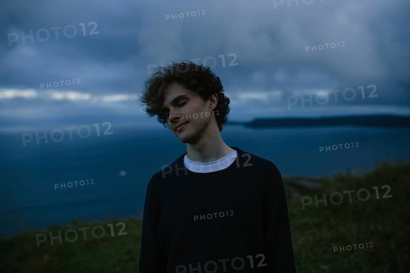 A thoughtful young man with eyes closed stands by the ocean in Signal Hill in St. John's, Newfoundland & Labrador, Canada. And he is lost in contemplation against a dramatic, cloudy seascape