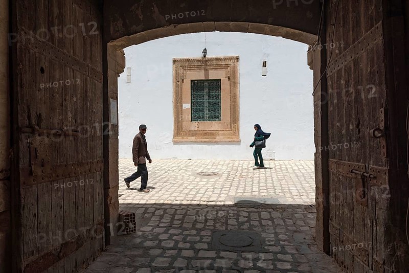 Tunisian men and women walk past some old wooden doors beside Bab Diwan the gate to Sfax Medina, Tunisia