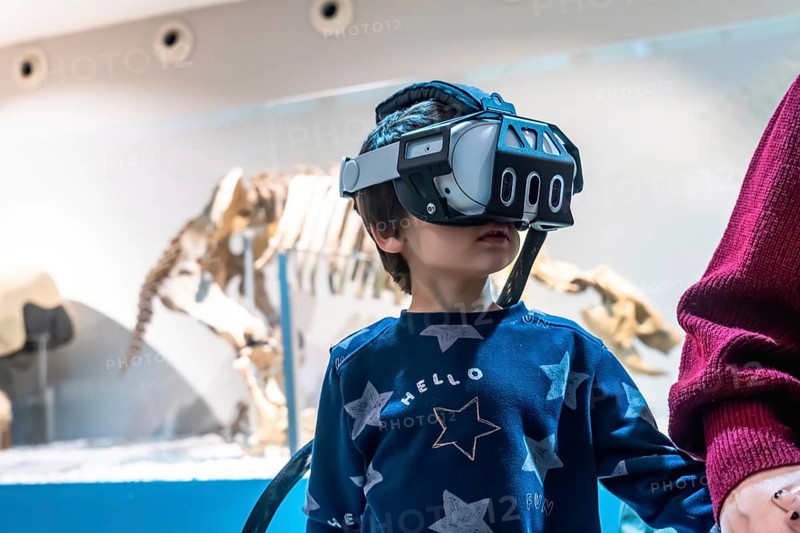 Young boy wearing a virtual reality headset, exploring an immersive experience at a museum with a dinosaur skeleton exhibit in the background, combining technology with education