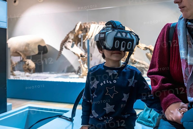 Young boy wearing a modern vr headset. Experiencing an immersive educational simulation at a museum. With dinosaur skeletons displayed in the background. Showcasing new interactive learning technology