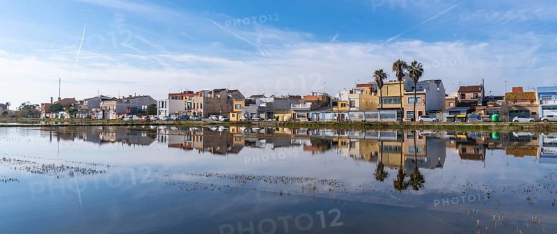 Traditional houses of el palmar fishing village reflecting perfectly on the calm water surface of an albufera rice field, showcasing spanish rural landscape and architecture under a blue sky