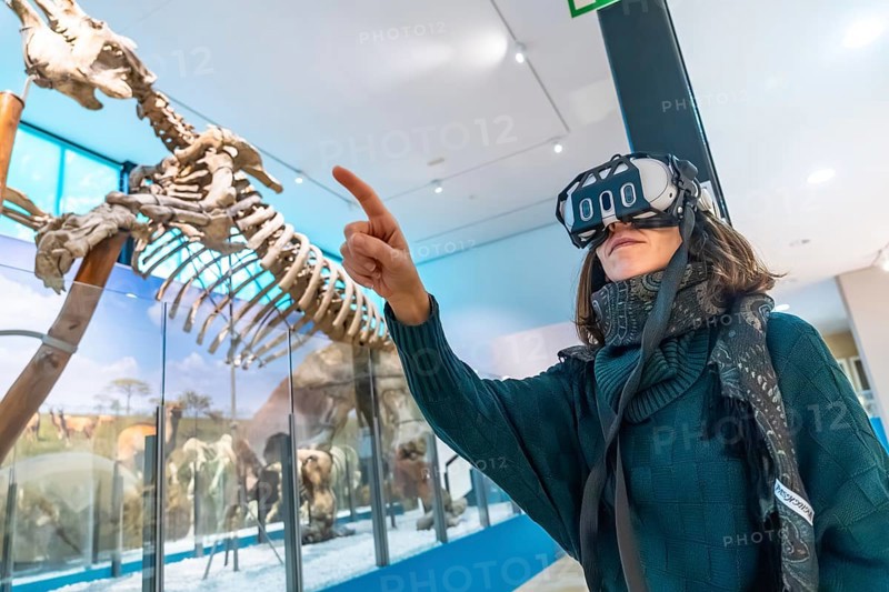 Woman wearing vr headset gestures and points at a life size dinosaur skeleton in a modern natural history museum, immersed in an educational mixed reality experience