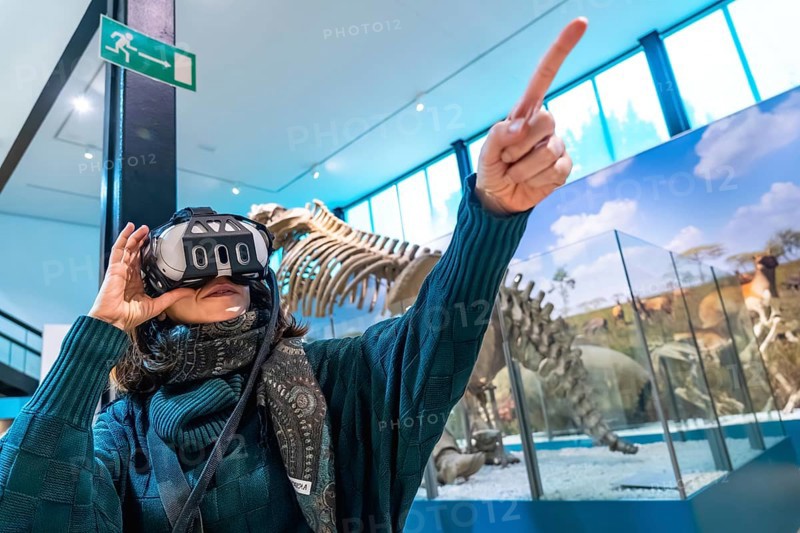 Woman wearing a virtual reality headset and pointing upward, interacting with an immersive educational experience featuring dinosaur skeletons and animal exhibits in a modern museum