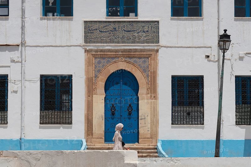 A Tunisian woman walks past the beautiful blue ornate door and architecture of the Abbasid School inside the Medina of Sfax, Tunisia, North Africa