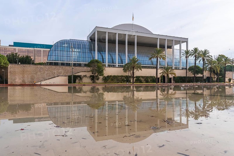 The palau de la musica music hall in valencia, spain, reflecting in a large permanent water feature or puddle with palm trees along the facade under a cloudy sky