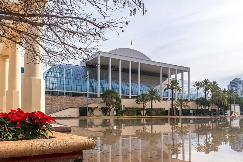 Palau de la musica, a modern concert hall featuring a glass facade and white columns, reflecting on a calm water pond surrounded by palm trees in turia gardens, valencia, spain