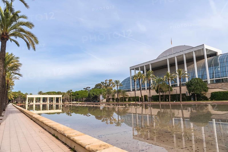 Palau de la musica building, an iconic landmark in valencia, reflecting in the turia gardens' calm waters surrounded by palm trees under a bright blue sky
