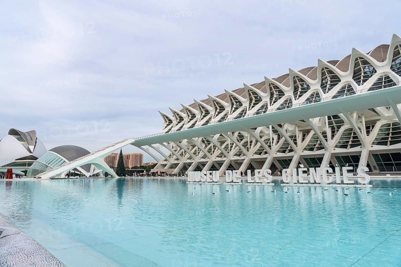 The museu de les ciencies principle building stands prominently against a cloudy sky, reflecting in the large light blue pool, showcasing its distinctive futuristic architecture
