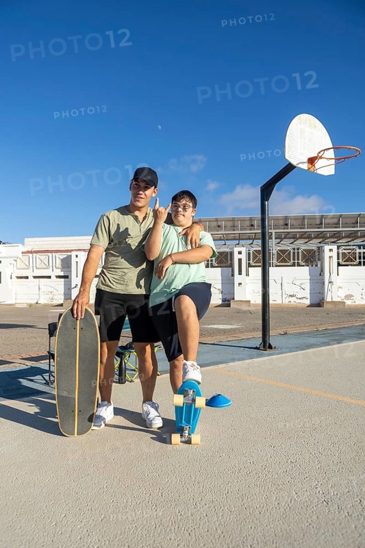 A teenager with Down Syndrome poses confidently with his brother and skateboards on an outdoor court, expressing pride, inclusion, and joy in a sunny and vibrant setting