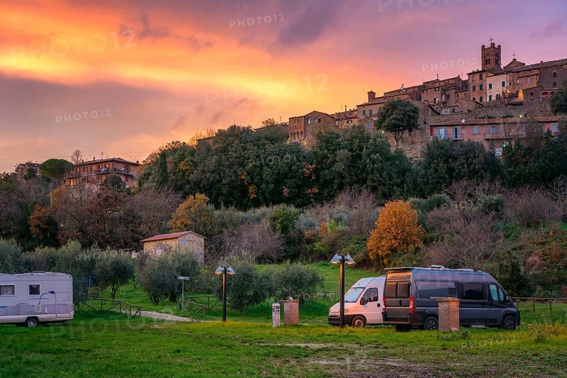 Camper vans and motorhomes parked in a scenic service area below a traditional Tuscany village. The hilltop stone houses and vibrant sunset sky create a picturesque countryside setting