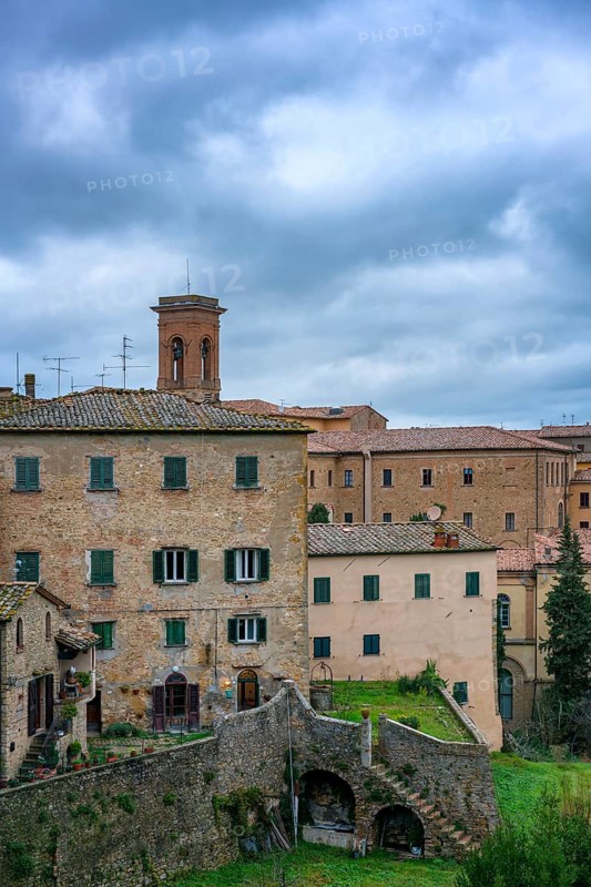 A charming view of Volterra, Tuscany, featuring traditional stone houses and rustic architecture. The medieval village is set against a dramatic cloudy sky, showcasing Italy's rich history