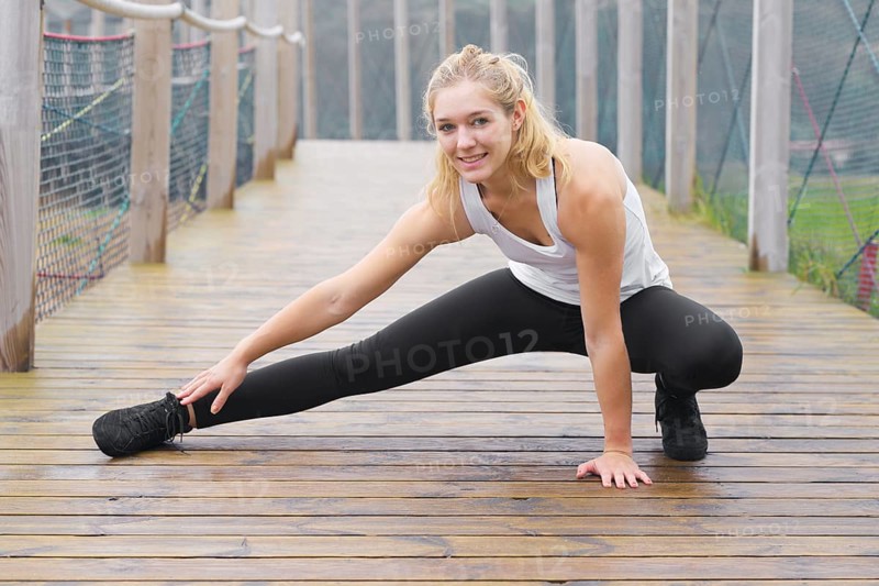 A young blond woman performs stretching exercises on a foggy wooden bridge, wearing sportswear and a focused expression