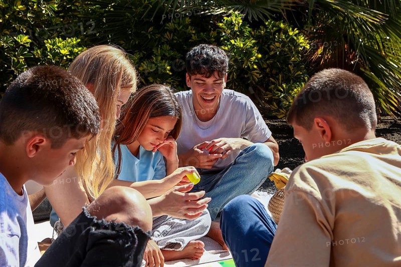 An older brother and sister enjoy time with their younger triplet siblings, two boys and a girl, in a sunlit garden at home, sharing smiles and a game