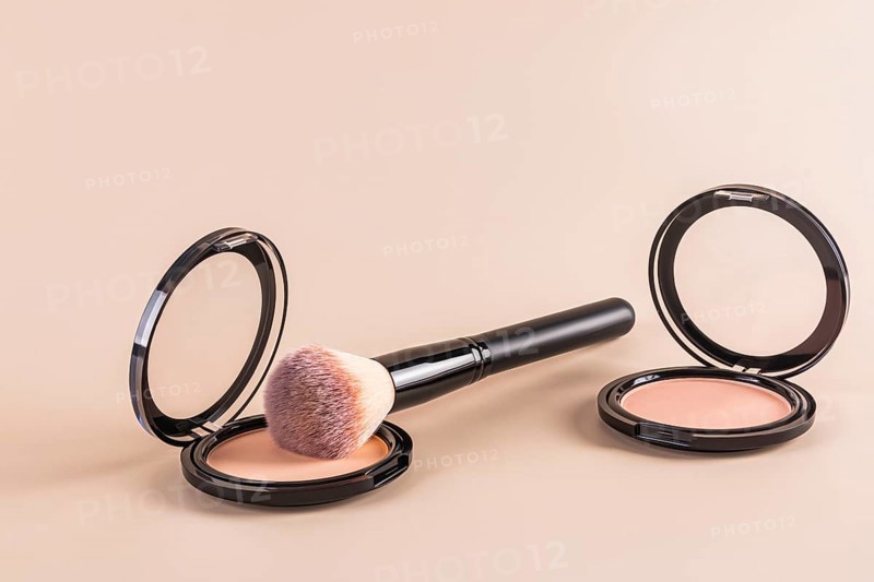 Luxurious black compact powder and a fluffy makeup brush beside an open loose powder container, all set against a muted beige backdrop. This setup highlights essential beauty tools for a flawless finish