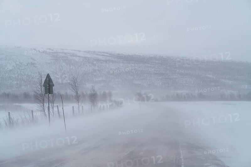 A blizzard envelops a remote road in Swedish Lapland, obscuring distant snow-covered hills and roadside fences. The winter landscape is stark and atmospheric
