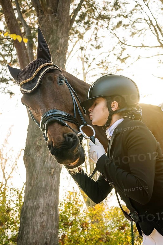 From below teenage girl, dressed in classical dressage attire, is affectionately interacting with a brown horse. She is closing her eyes and about to kiss the horse. The scene is illuminated by soft, natural light, creating a serene atmosphere