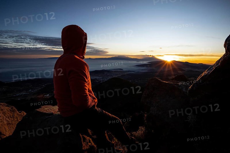 Back view of an unrecognizable man sitting on a mountain peak, watching a stunning sunrise over a cloudy landscape