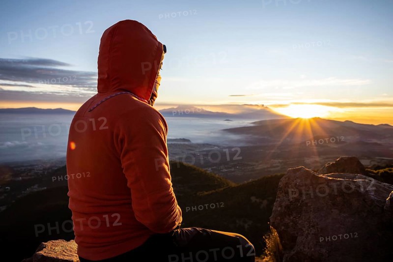 Back view of unrecognizable man sitting atop a mountain, watching the sunrise. The early morning light casts warm hues over the landscape, illuminating the distant mountains and clouds