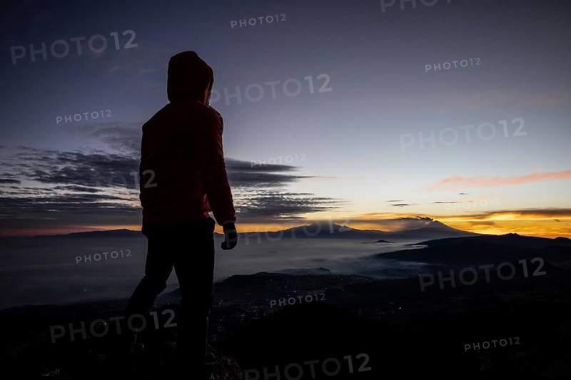 Back view of unrecognizable man standing at the summit during sunrise, overlooking distant mountains and clouds, with vibrant sky colors in the background