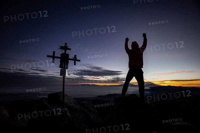 Unrecognizable man stands with arms raised near a silhouetted cross at sunrise, atop a rugged mountain peak, highlighting a moment of triumph and peace
