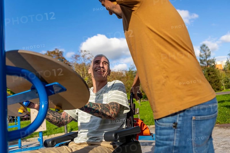 Friends enjoy a sunny day in the park, focusing on inclusive fitness for cerebral palsy One is using a wheelchair-accessible exercise machine, smiling, and interacting with a standing friend