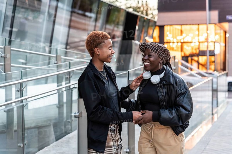 Happy LGBT multiethnic couple enjoying a cheerful moment outdoors in the city during sunset, dressed stylishly in casual attire, with one partner wearing headphones