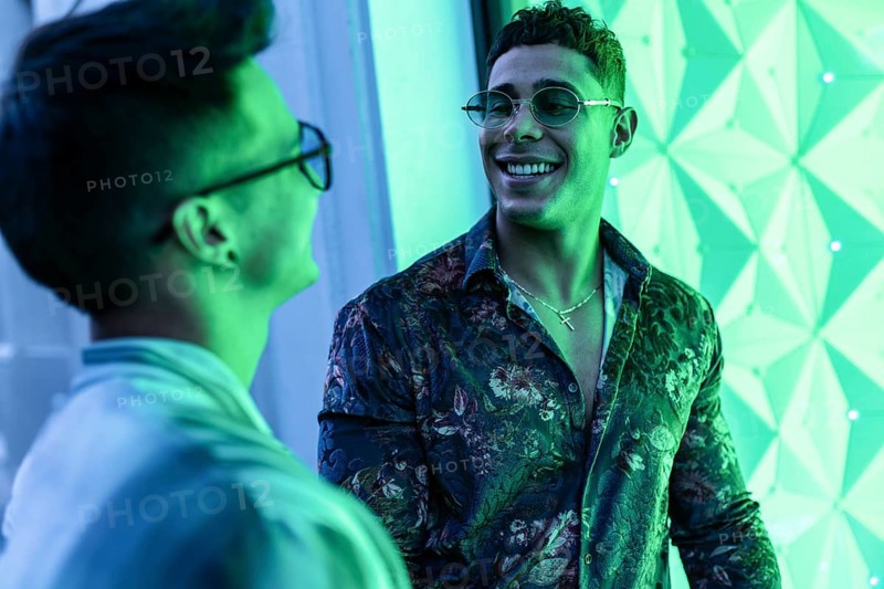 Two latin gay men smiling warmly at each other in an illuminated urban setting. Vibrant green tones create a lively atmosphere, highlighting their joyful connection and stylish attire