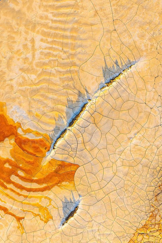 Aerial view of cracked mud in vibrant hues, showcasing an abstract pattern of orange, blue, and beige The intricate design highlights the beauty of natural textures