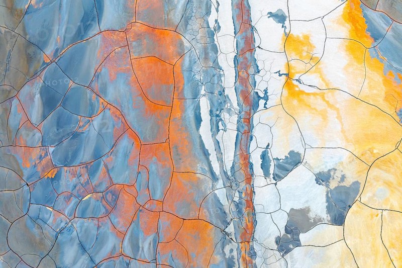 Aerial view of multicolored cracked mud showing vibrant hues of blue, orange, and yellow This abstract natural pattern resembles a dynamic, artistic painting