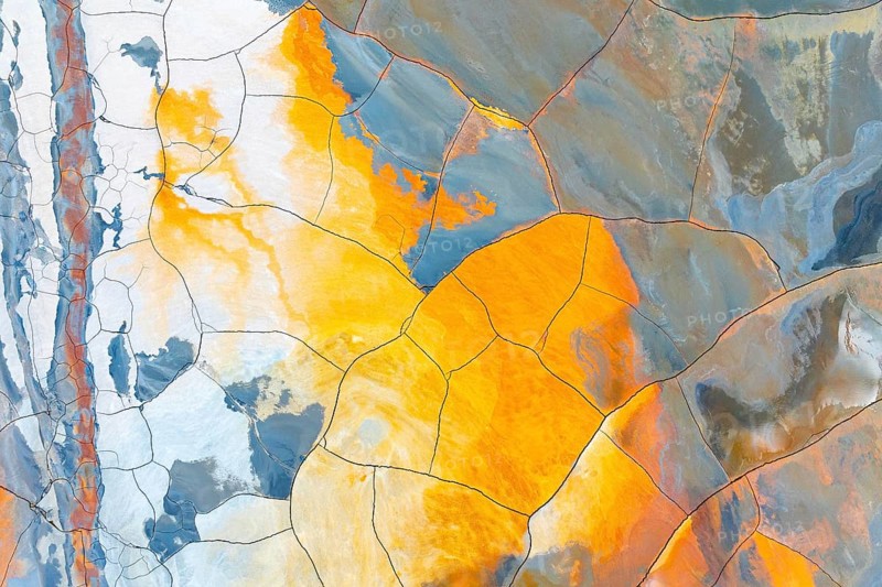 Aerial view of multicolored cracked mud, featuring vibrant hues of orange, blue, and white The intricate patterns form a unique, natural abstract artwork