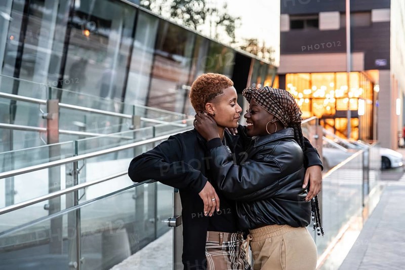 An affectionate LGBT multiethnic couple embraces outdoors in the city, capturing a candid and heartfelt moment. They are surrounded by modern buildings and soft evening lighting