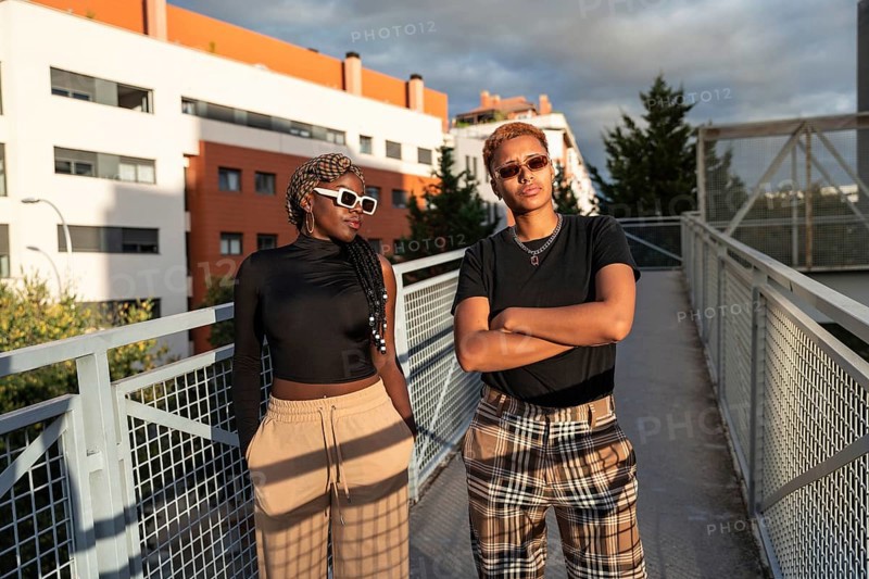 A stylish LGBT multiethnic couple poses confidently on an urban bridge under the warm, golden sunlight. Both wear sunglasses and casual attire, expressing joy and togetherness