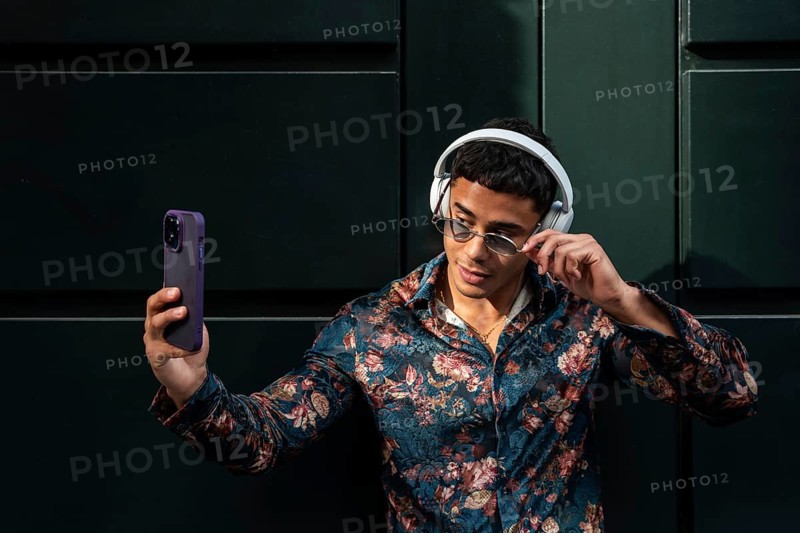 A trendy latin man in floral shirt and sunglasses takes a selfie outdoors, showcasing modern style with headphones on. The photo captures confidence and contemporary fashion