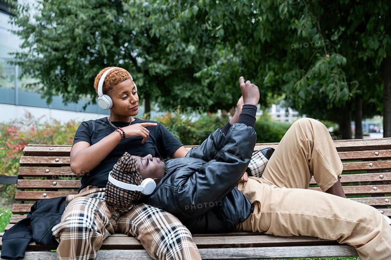 A multiethnic LGBT couple enjoying a peaceful moment on a park bench, each wearing headphones. One person is lying down, affectionately looking at the other, surrounded by lush greenery