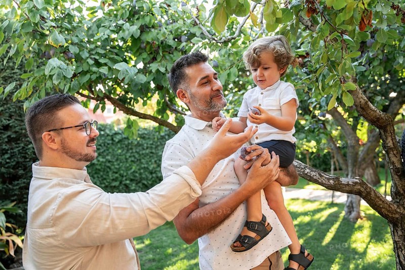 A joyful scene of a gay couple spending quality time with their young son outdoors. They are in a lush garden, symbolizing love, family bonding, and inclusivity