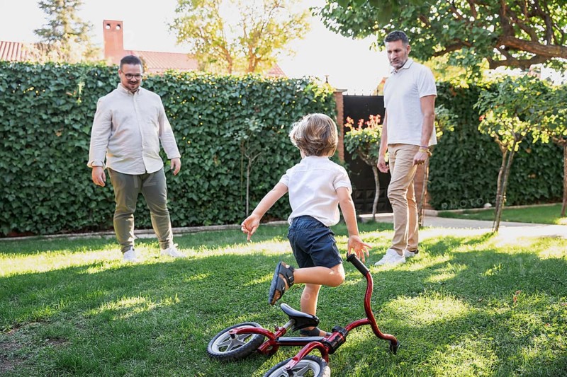 A loving gay couple shares a joyful moment with their young son playing with bicycle in the garden. The sunlight creates a warm atmosphere, highlighting their family bond