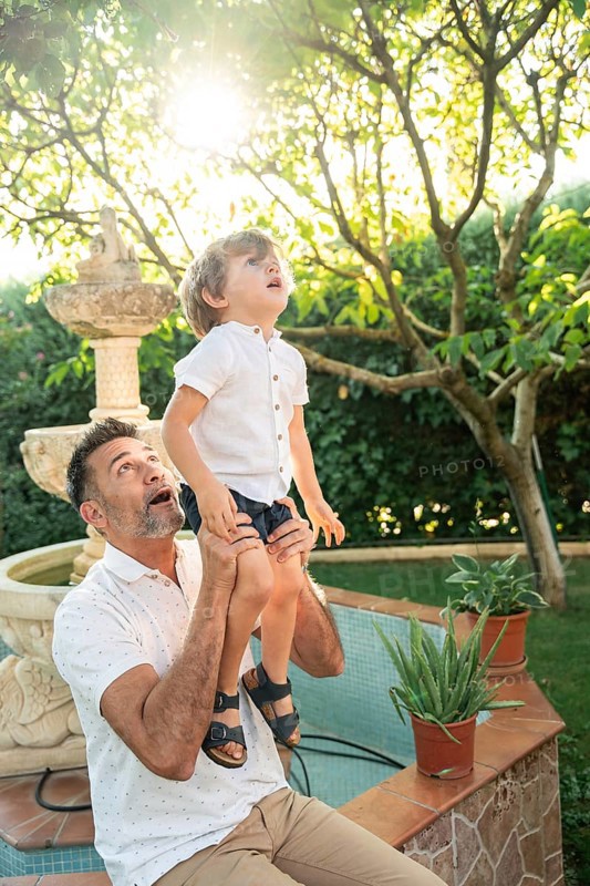 A father lifts his son as they gaze at something fascinating. Sunlight filters through the leaves in a lush garden, creating a warm, serene atmosphere