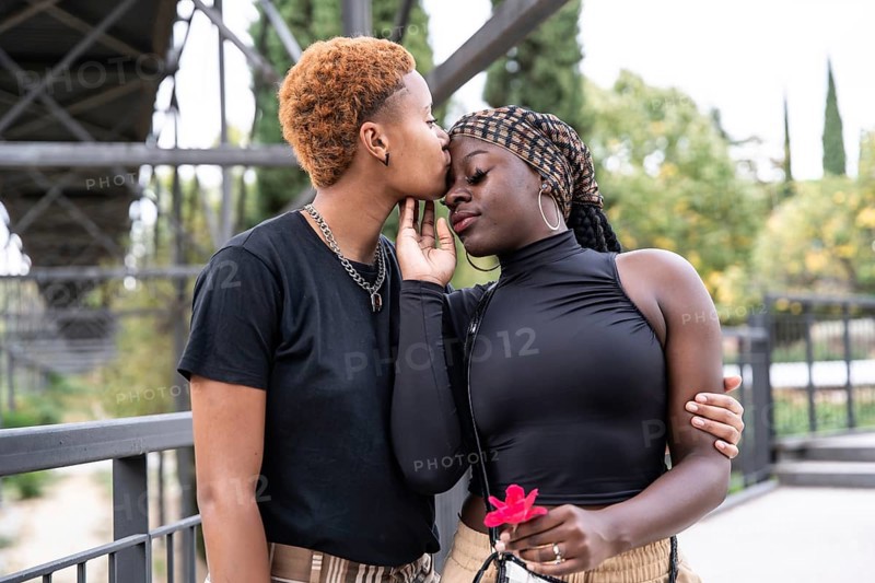 A loving LGBTQ multiethnic couple embraces in a park, sharing a tender moment. One partner gently kisses the other's forehead, exemplifying a genuine connection and affection