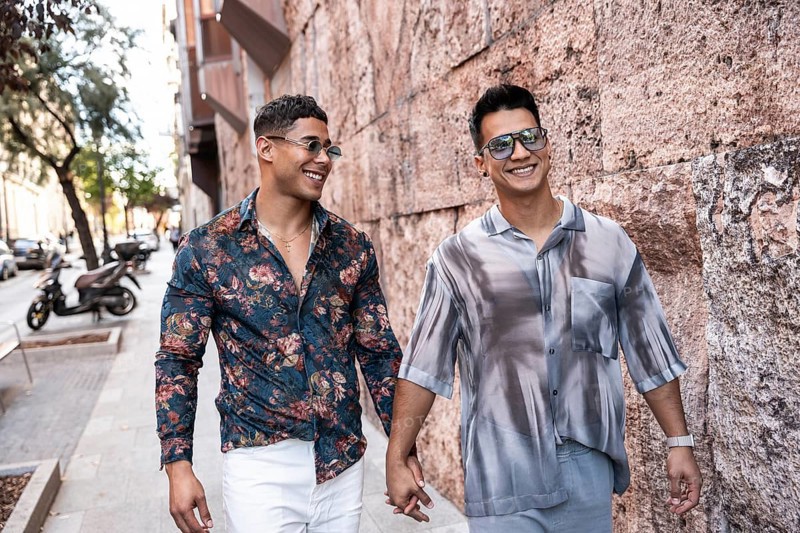 A joyful latin gay couple walks hand in hand down a lively street in Madrid. They're smiling, wearing stylish outfits and sunglasses, enjoying the sunny day and vibrant city atmosphere