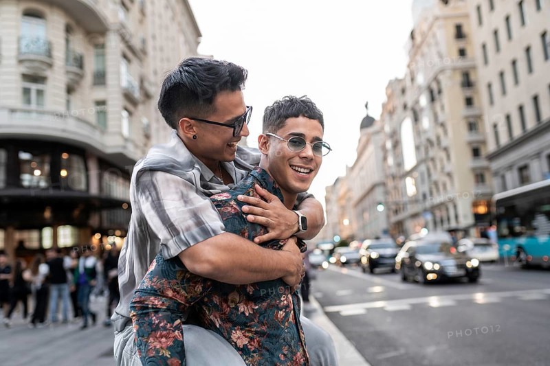 A latin gay couple shares a joyful moment on a bustling street in Madrid, surrounded by beautiful architecture and city life. They express happiness and love in this urban setting