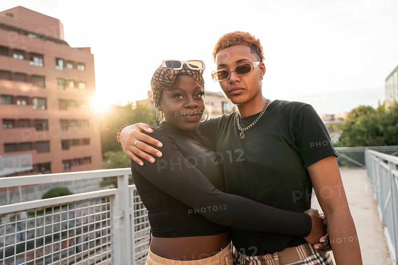 A loving LGBT multiethnic couple embraces outdoors on a bridge during a beautiful sunset. The warm light enhances their affectionate pose, symbolizing unity and love in an urban setting